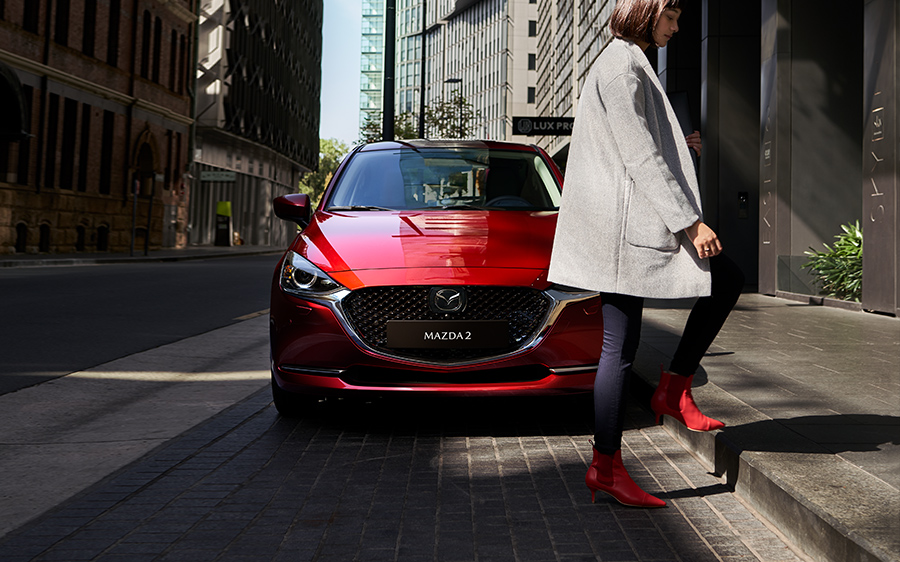 Mazda2 Modellabbildung in der Modellfarbe rot in Frontansicht mit vorbeigehender Frau